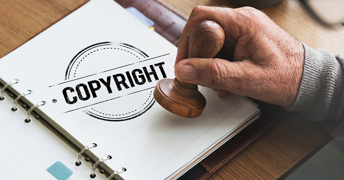 A hand holding a wooden stamp over a document with a "Copyright" mark, symbolizing intellectual property protection.