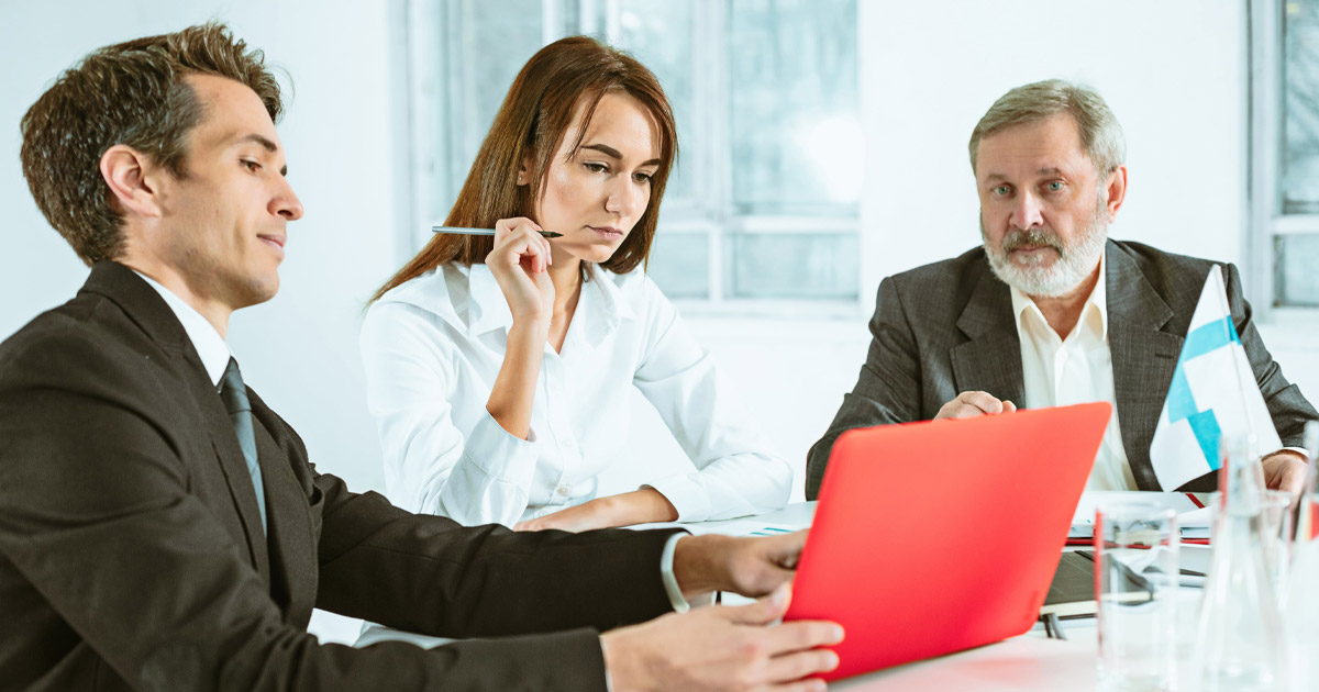 Business professionals in a meeting, reviewing documents on a red laptop in a bright office setting.