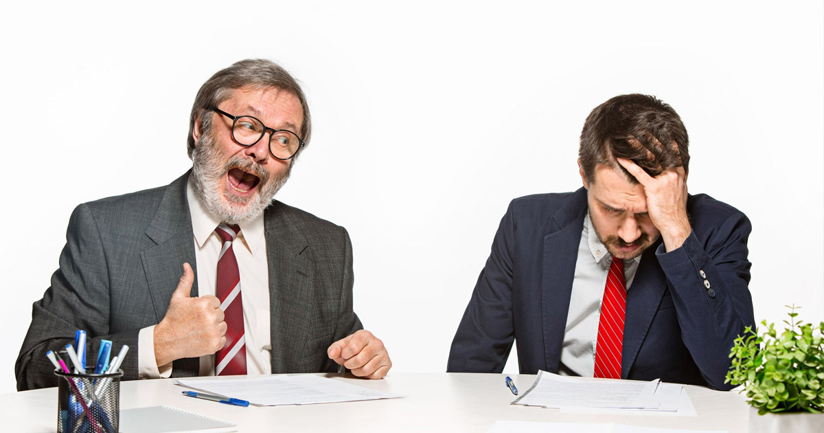 Two businessmen at a desk, one excited and giving thumbs up, the other stressed with head in hand during a meeting