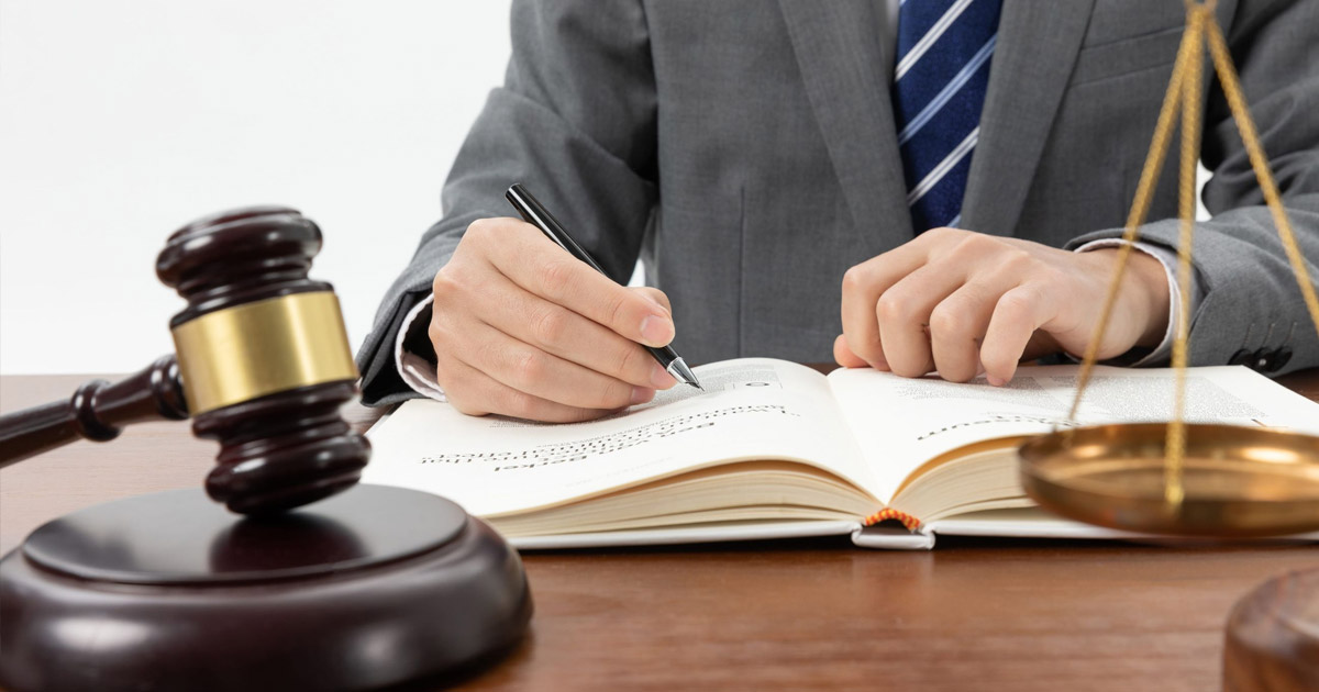 Legal professional writing in law book with gavel and justice scales, wearing gray suit and blue striped tie