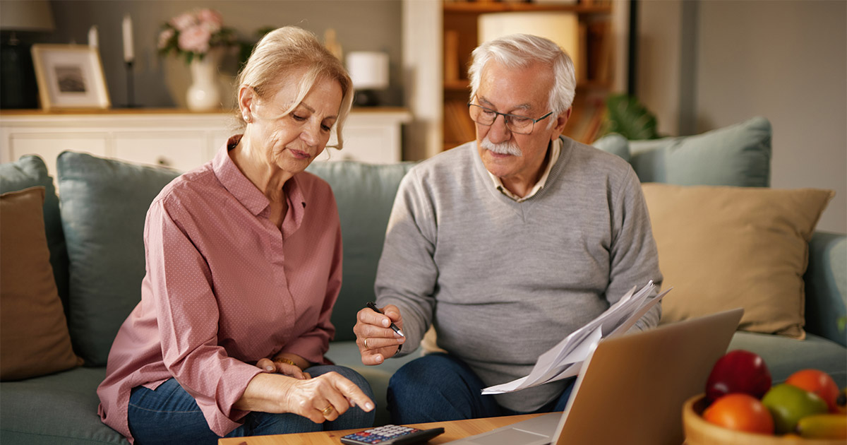 Retired couple reviewing finances together at home, using laptop and calculator on cozy living room couch