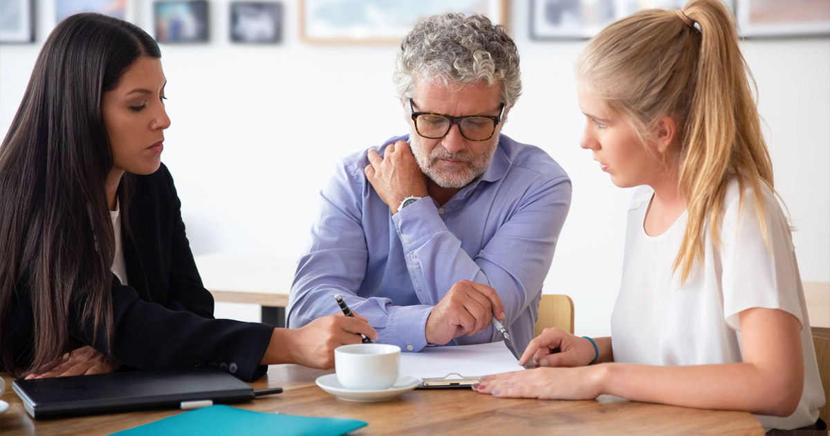 Three people in a meeting, older professional man reviewing document with two women at table with coffee cup