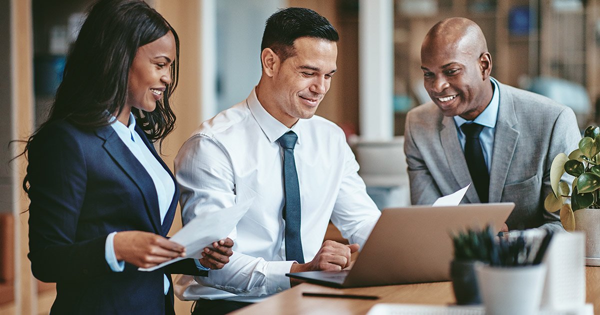 Three business professionals reviewing documents and collaborating over a laptop in a modern office setting, smiling and engaged.