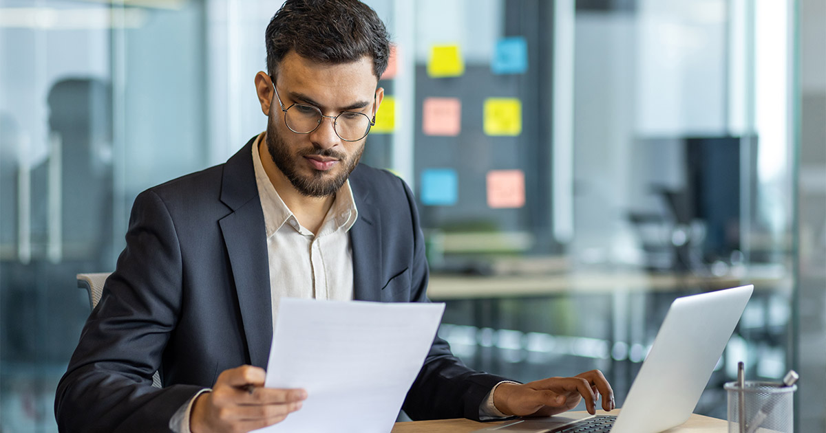 Professional in business attire working on laptop and reviewing document in modern office with sticky notes in background